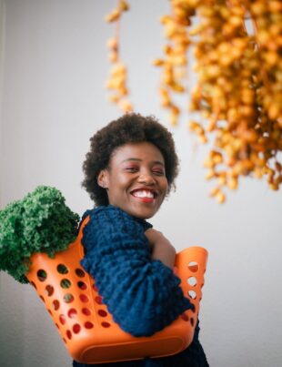 happy young black woman holding basket with lettuce on shoulder and cluster of yellow dates in grocery store on gray background