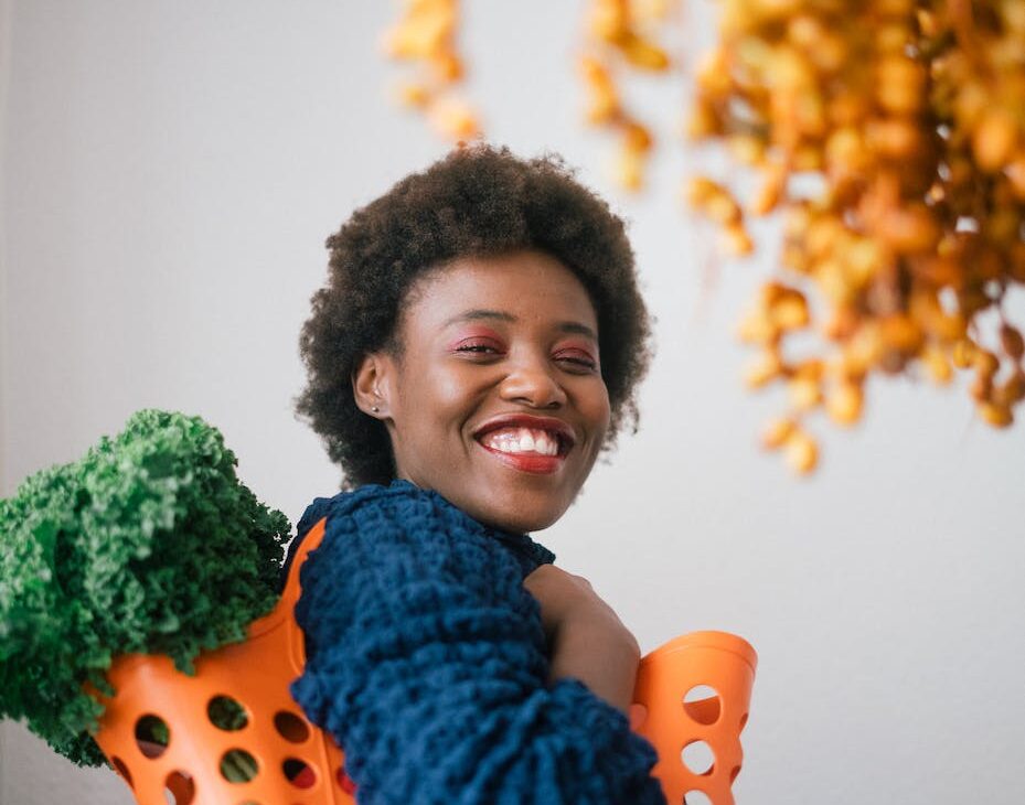 happy young black woman holding basket with lettuce on shoulder and cluster of yellow dates in grocery store on gray background