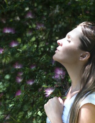 woman closing her eyes against sun light standing near purple petaled flower plant