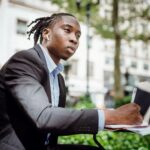 thoughtful black man writing in notebook sitting at table in outdoor cafeteria