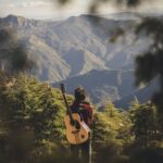 person with acoustic guitar standing in green field near mountain