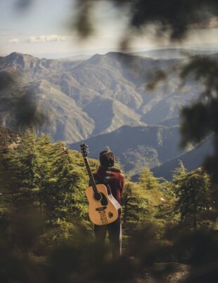 person with acoustic guitar standing in green field near mountain