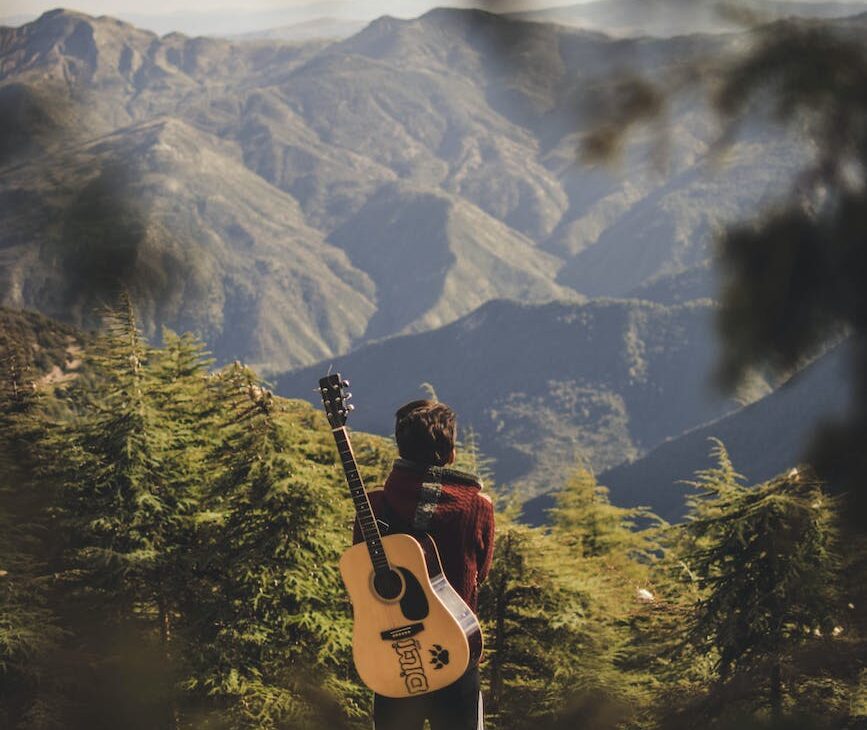 person with acoustic guitar standing in green field near mountain